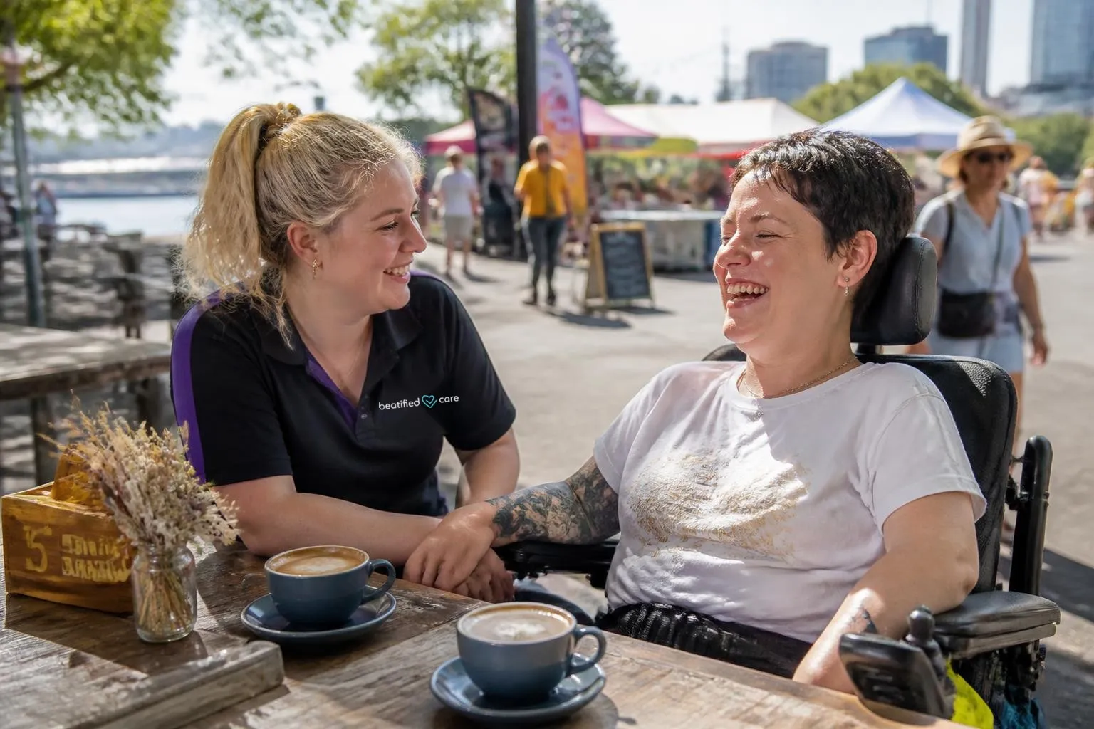 Woman using a wheelchair laughing with her support worker at a riverside cafe